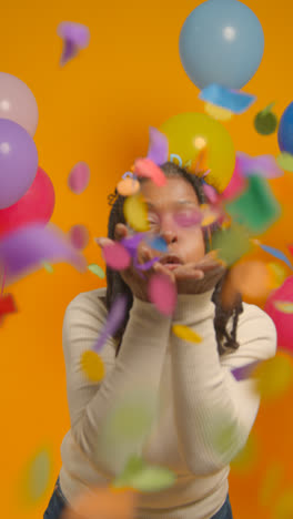 Vertical-Video-Studio-Portrait-Of-Woman-Wearing-Birthday-Headband-Celebrating-Blowing-Paper-Party-Confetti-To-Camera